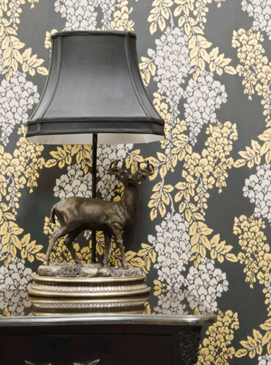 壁：FARROW&BALL 壁紙 2206 Wisteria / 小さなチェストの背景に黒ベースに白や黄色の藤の花が施された壁紙。チェストの上には黒のシェードと馬のアートが飾られたランプ。