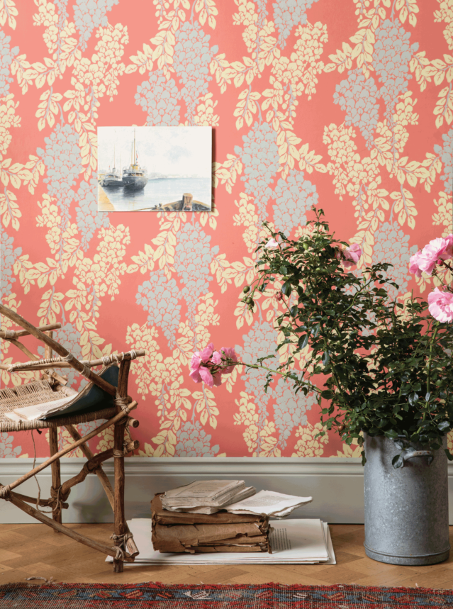 壁：FARROW&BALL 壁紙 2222 Wisteria / インテリアの一角を藤の花モチーフになっている壁紙が貼られている。壁紙の地色はソフトな赤にグレーの藤の花とクリーム色の藤の葉が描かれている。巾木はFarrow&Ball No.18 French Grayで塗装されている。壁の前には植物やチェア、古い本が飾られている。