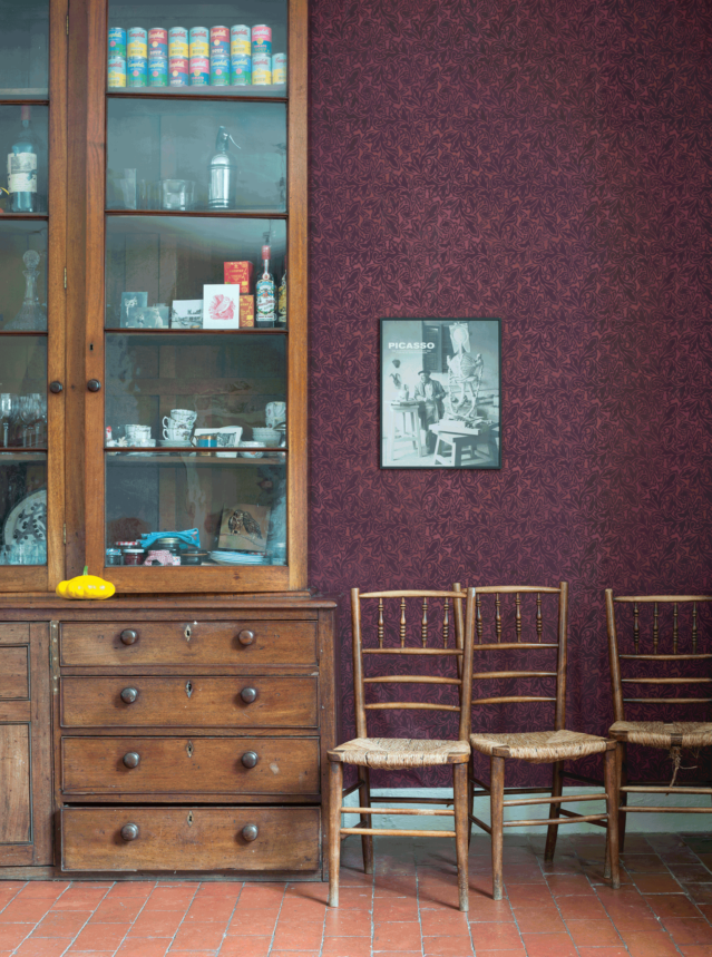 壁：FARROW&BALL 壁紙 4904 Feuille / 赤紫系統のベースカラーの上に、ダークカラーの植物柄が施されている壁があるキッチン。木製の背の大きな食器棚と木製のチェア３脚が置かれている。