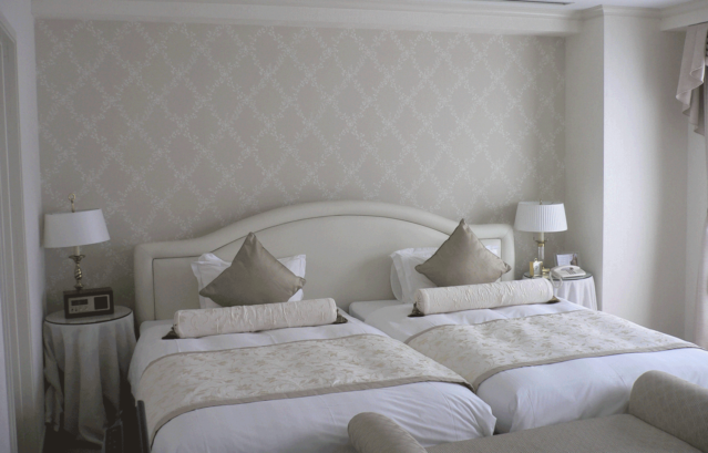 写真提供：北野ホテル / 壁：FARROW&BALL 壁紙 620 Toile Trellis / 神戸の老舗ホテルである北野ホテルの一室。寝室のホワイトのベッドの背景の壁に、淡く落ち着きのあるベージュのボタニカル柄の壁紙。